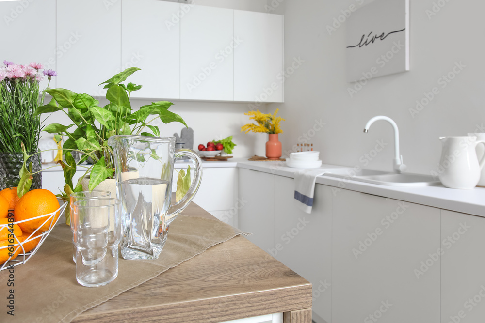 Jug of water, glasses and fruit basket on wooden island table in modern kitchen, closeup