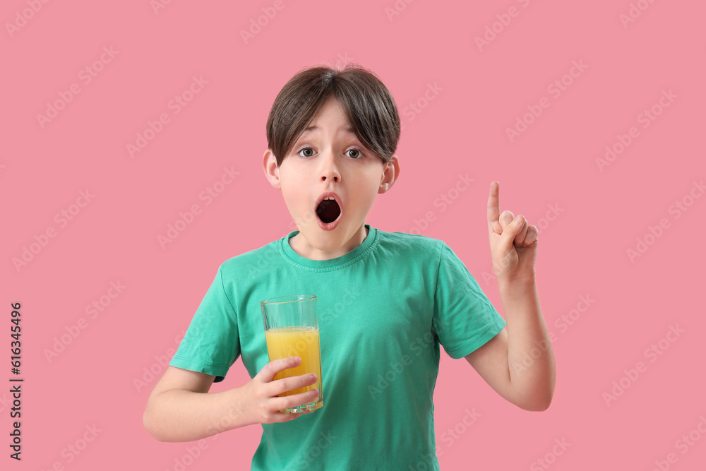 Shocked little boy with glass of orange juice pointing at something on pink background