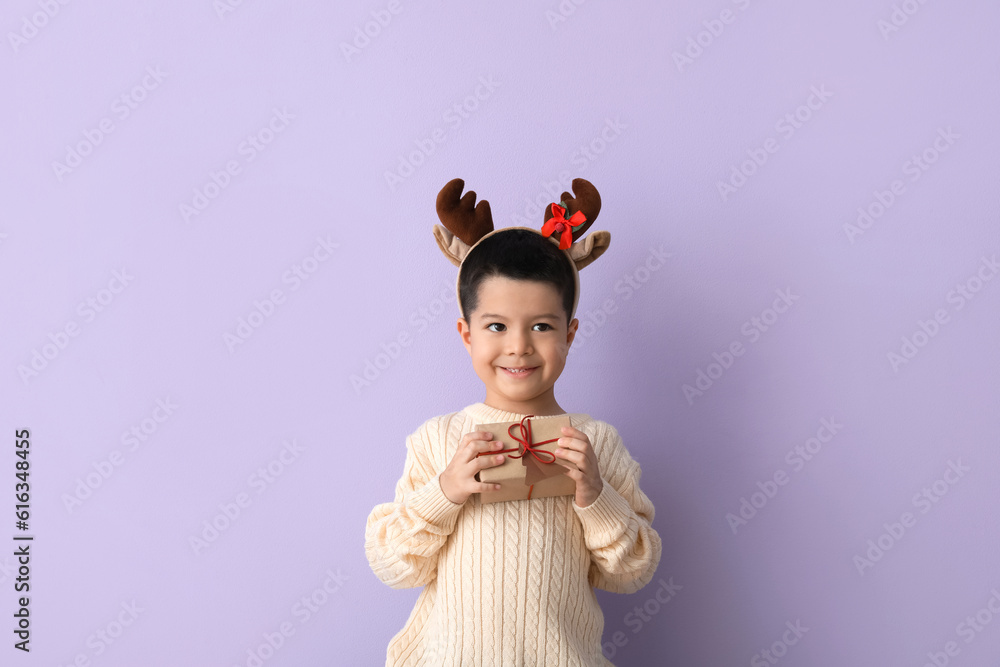 Cute little boy in deer horns with gift on lilac background