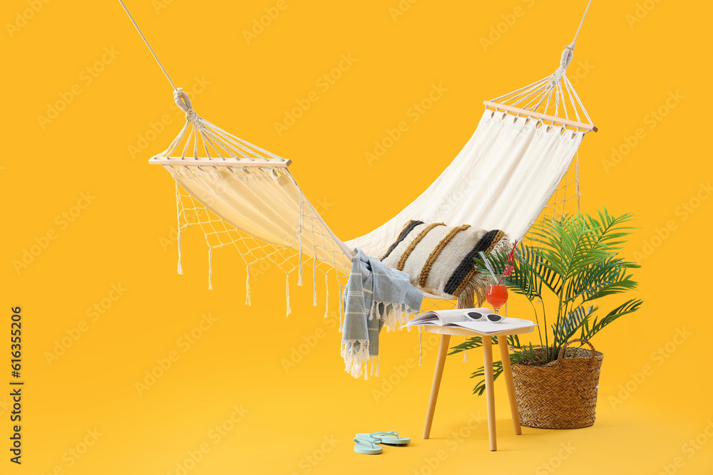 Cozy hammock with plant, beach accessories and cocktail on stool against yellow background. Summer v