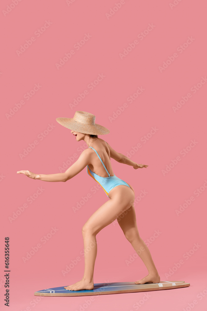 Young beautiful woman in swimsuit and wicker hat with surfboard on pink background