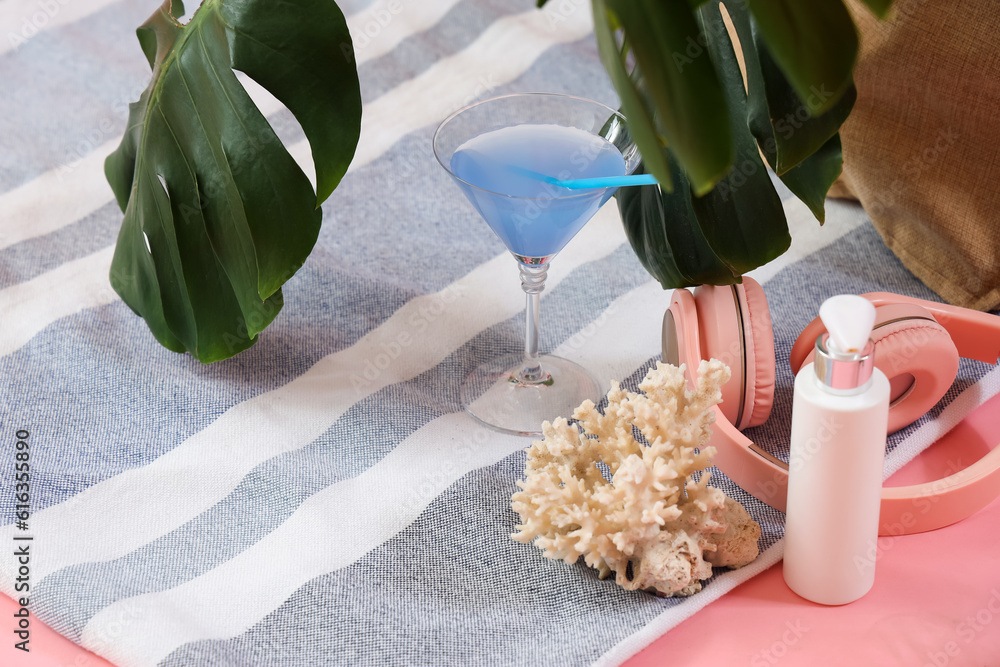 Cocktail with bottle of sunscreen cream, headphones and coral on beach blanket against pink backgrou