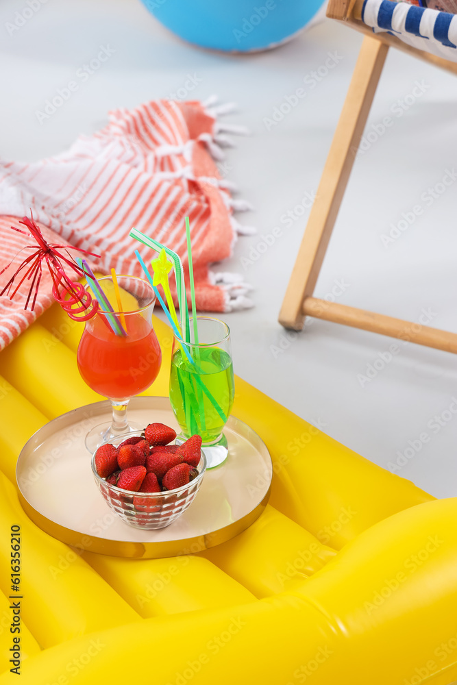 Cocktails with bowl of strawberry on inflatable mattress against grey background. Travel concept