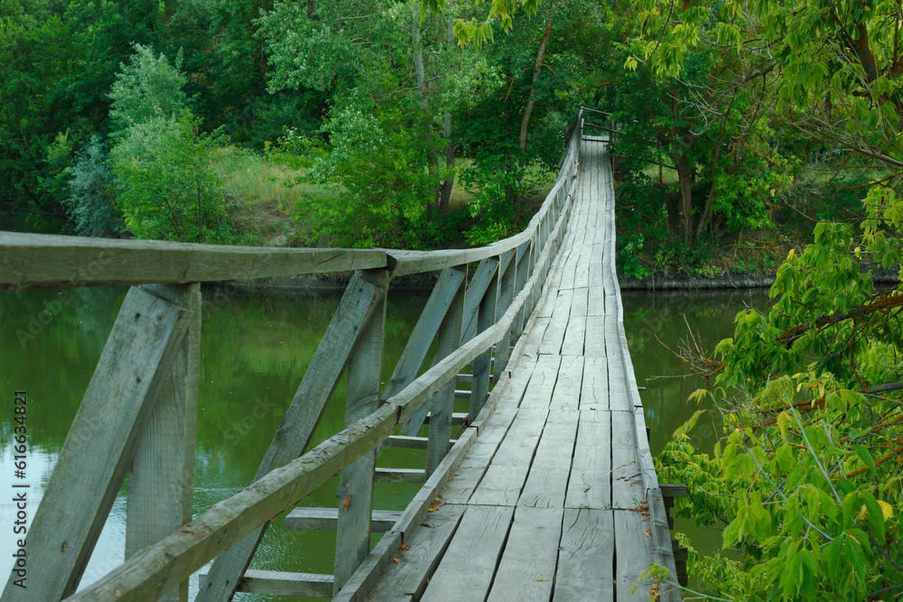 Suspension wooden bridge on river. Green trees on riverside.