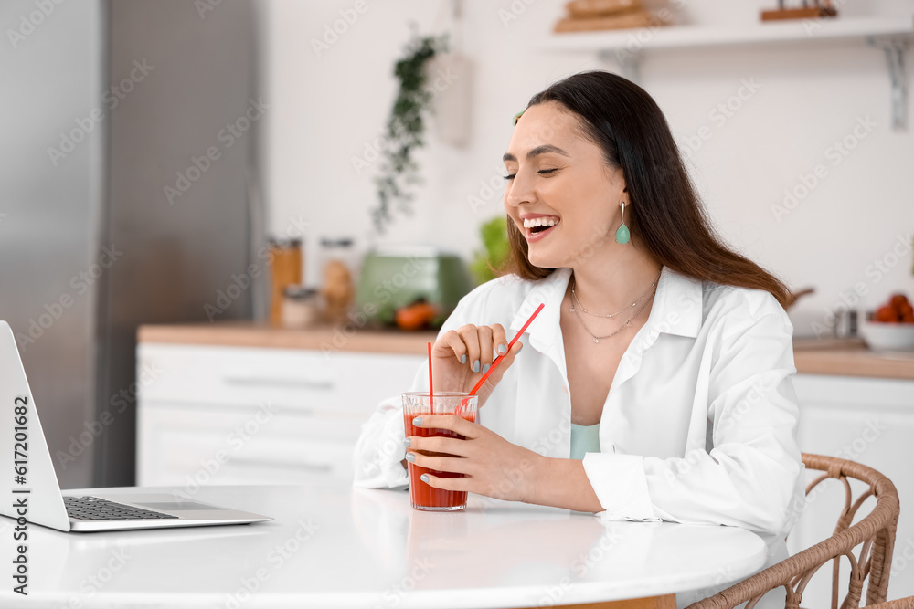 Young woman with glass of vegetable juice and laptop in kitchen