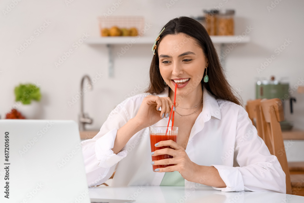 Young woman with glass of vegetable juice and laptop in kitchen