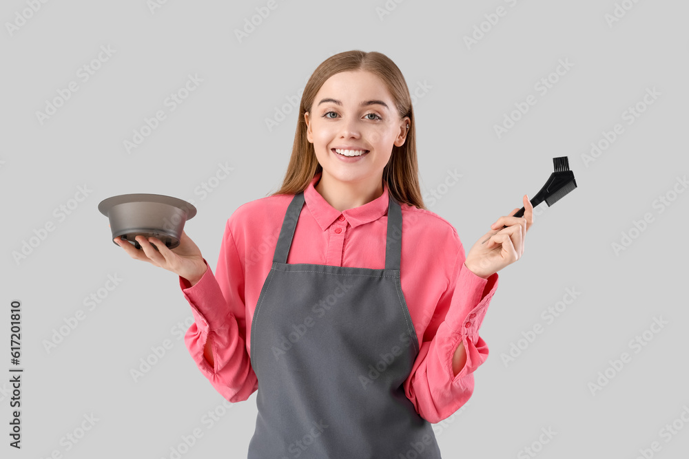 Female hairdresser with bowl and brush for hair dye on grey background