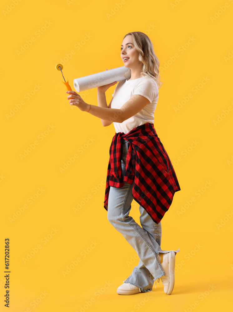 Young woman with wallpaper and roller on yellow background