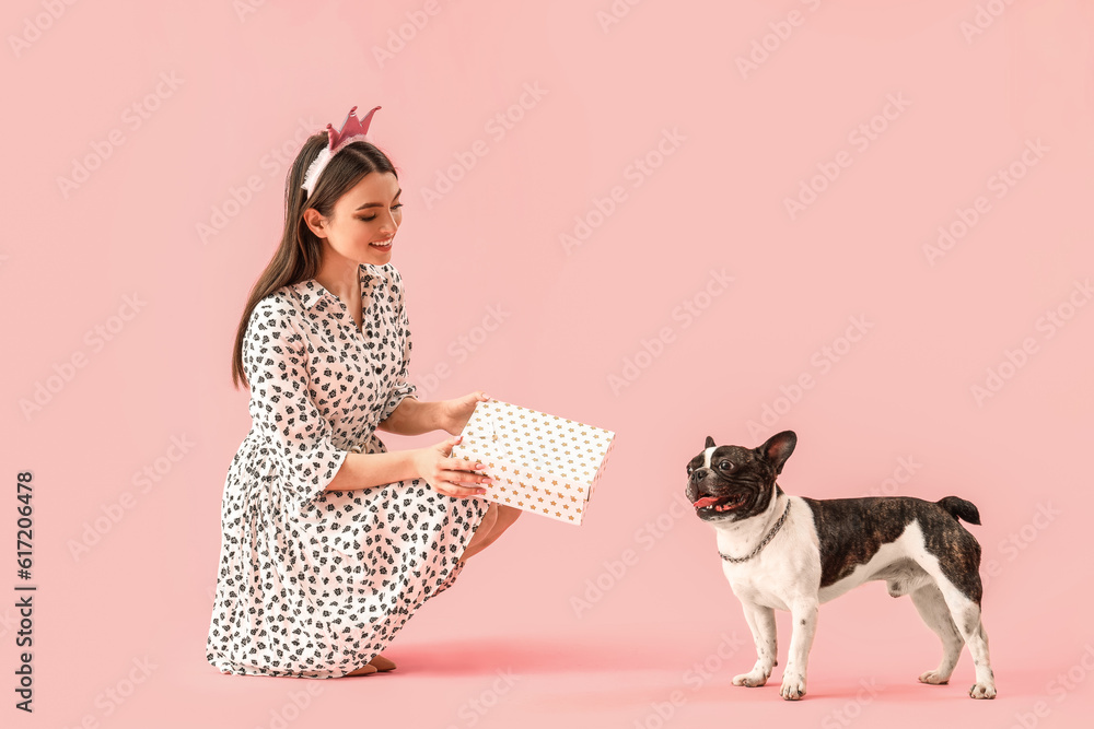 Young woman with birthday gift and her French bulldog on pink background