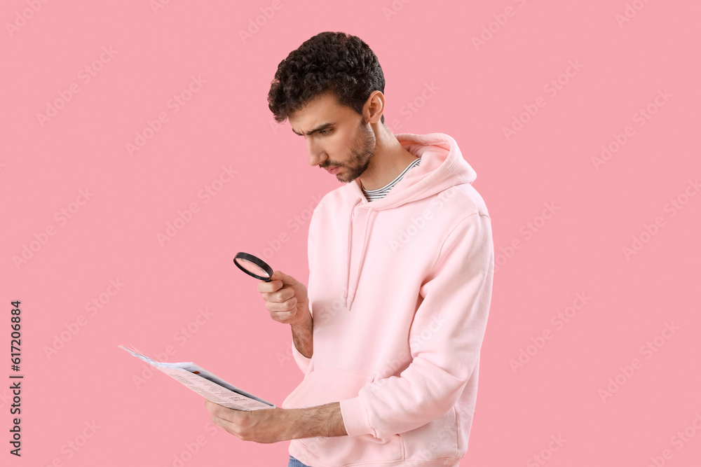 Young man with magnifier reading newspaper on pink background