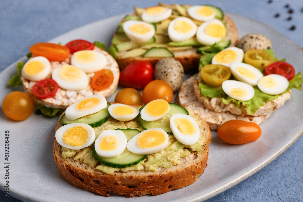 Different tasty toasts with quail eggs, avocado and tomatoes in plate on blue background, closeup