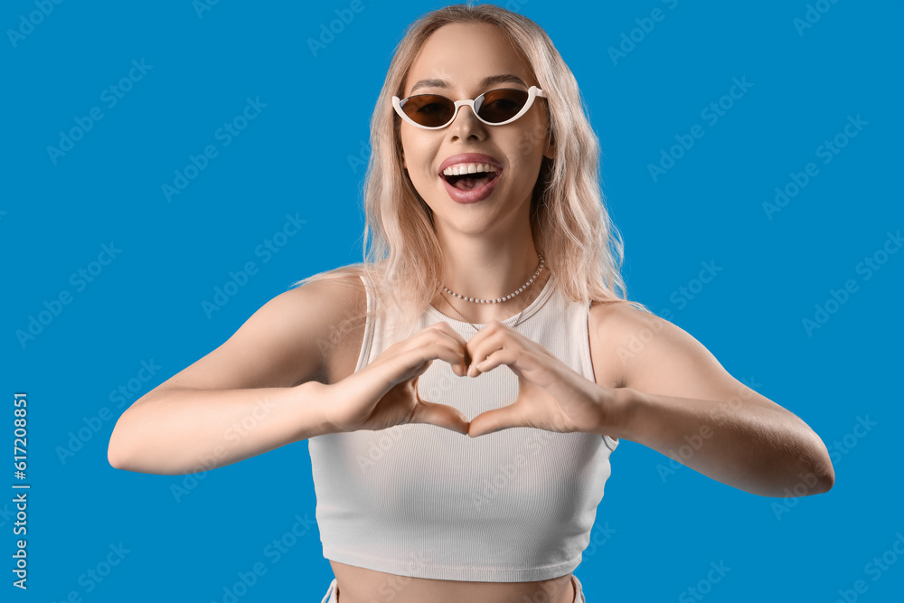 Young woman in stylish sunglasses making heart with her hands on blue background