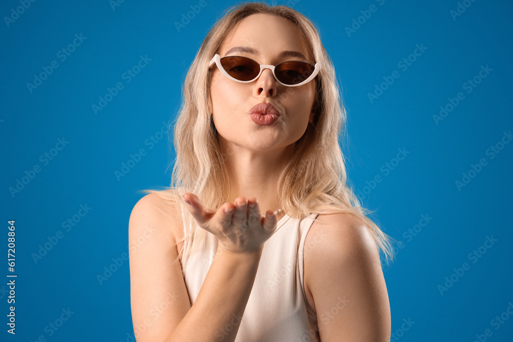 Young woman in stylish sunglasses blowing kiss on blue background, closeup