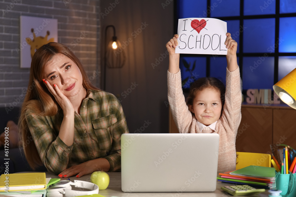 Little girl holding paper with text I DONT LOVE SCHOOL and her mother at home late in evening