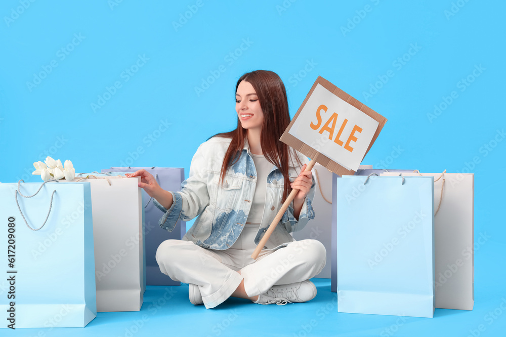 Young woman with sale sign and shopping bags sitting on blue background