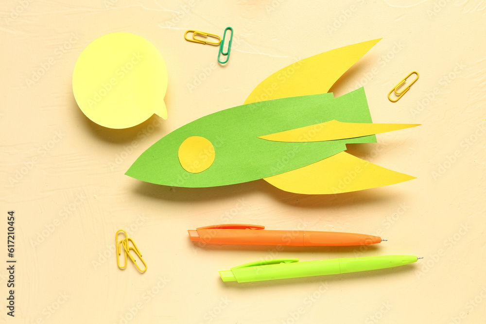 Green paper rocket with pens, sticker and clips on beige background