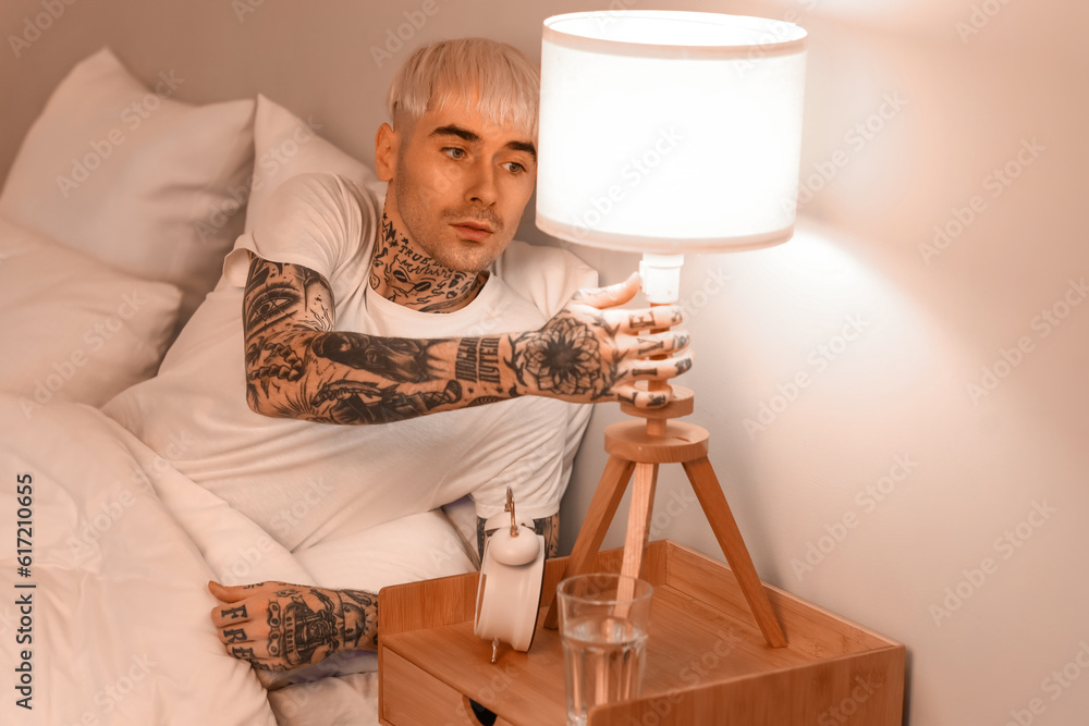 Young tattooed man turning off lamp in bed at night