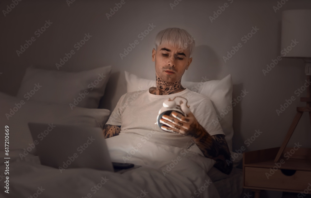 Young tattooed man with alarm clock and laptop in bed at night