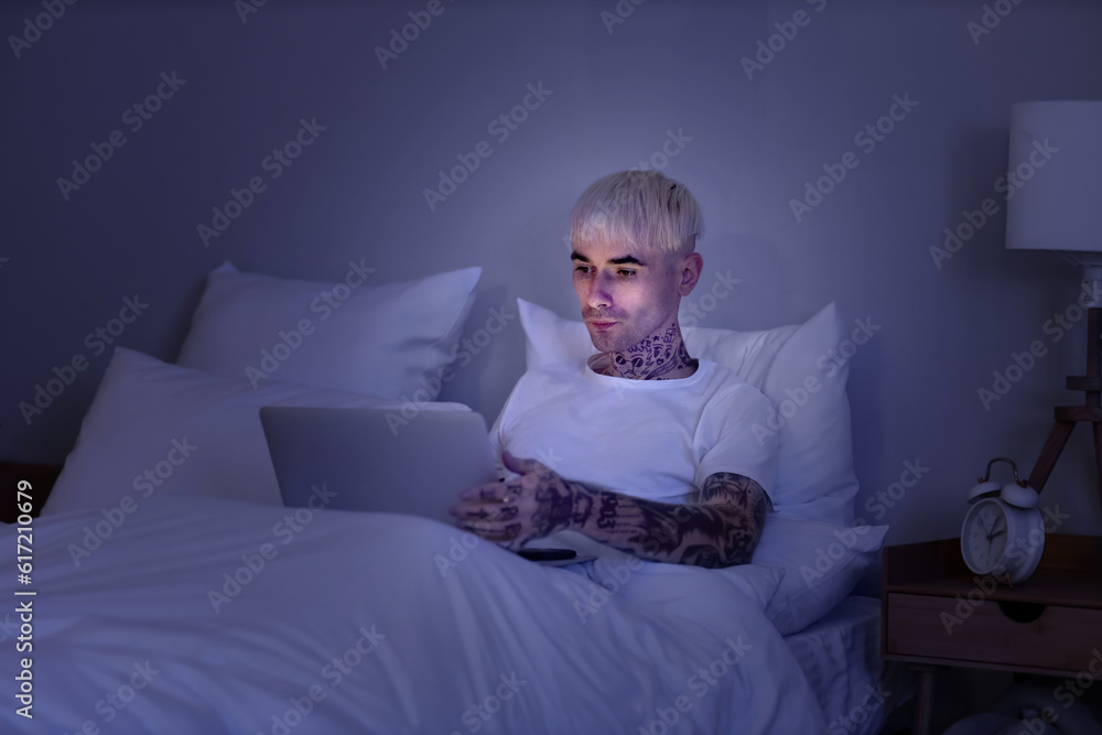 Young tattooed man using laptop in bed at night