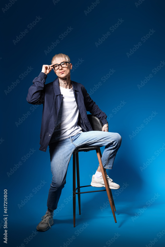 Young tattooed man in eyeglasses sitting on blue background