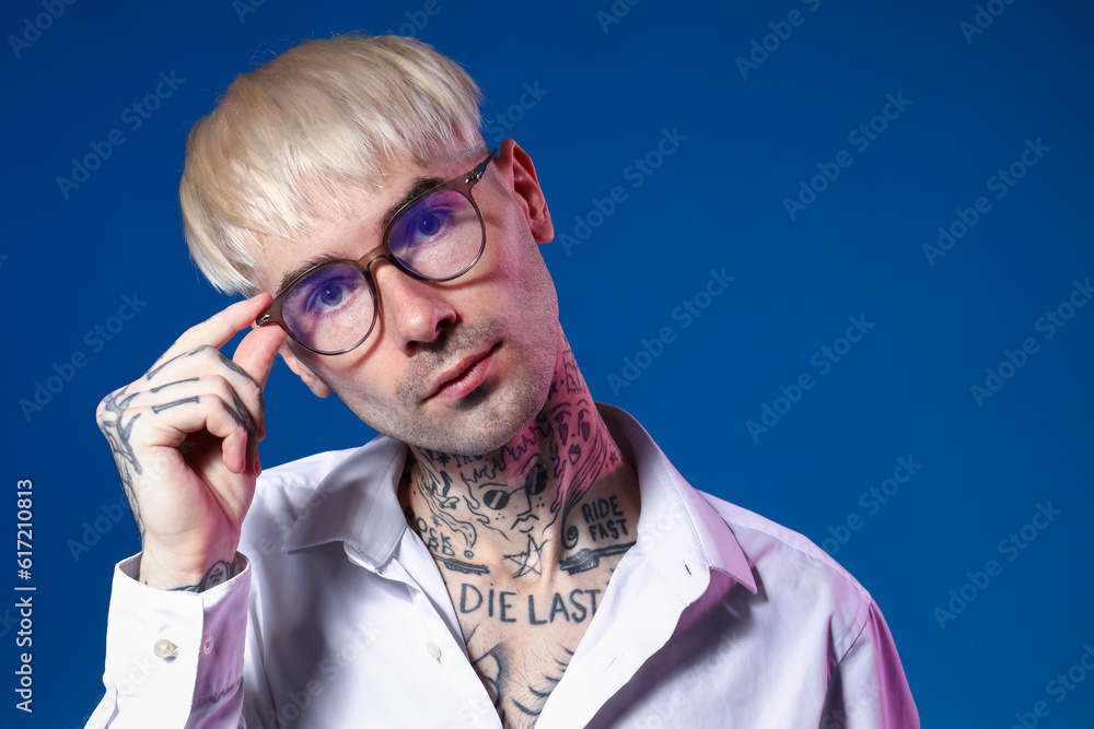 Young tattooed man in eyeglasses on blue background, closeup