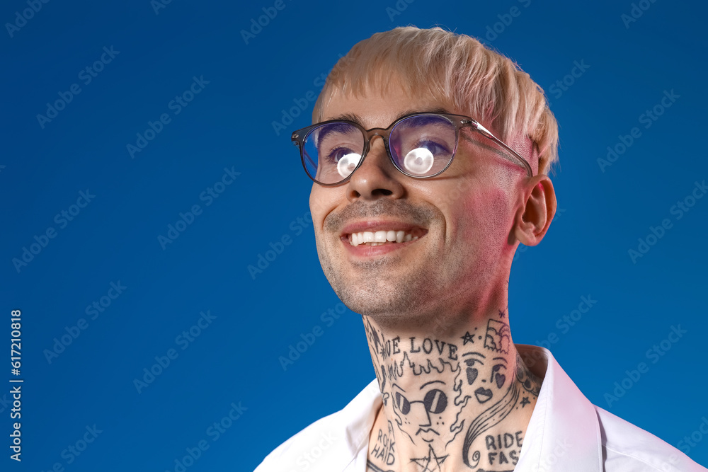 Young tattooed man in eyeglasses on blue background, closeup