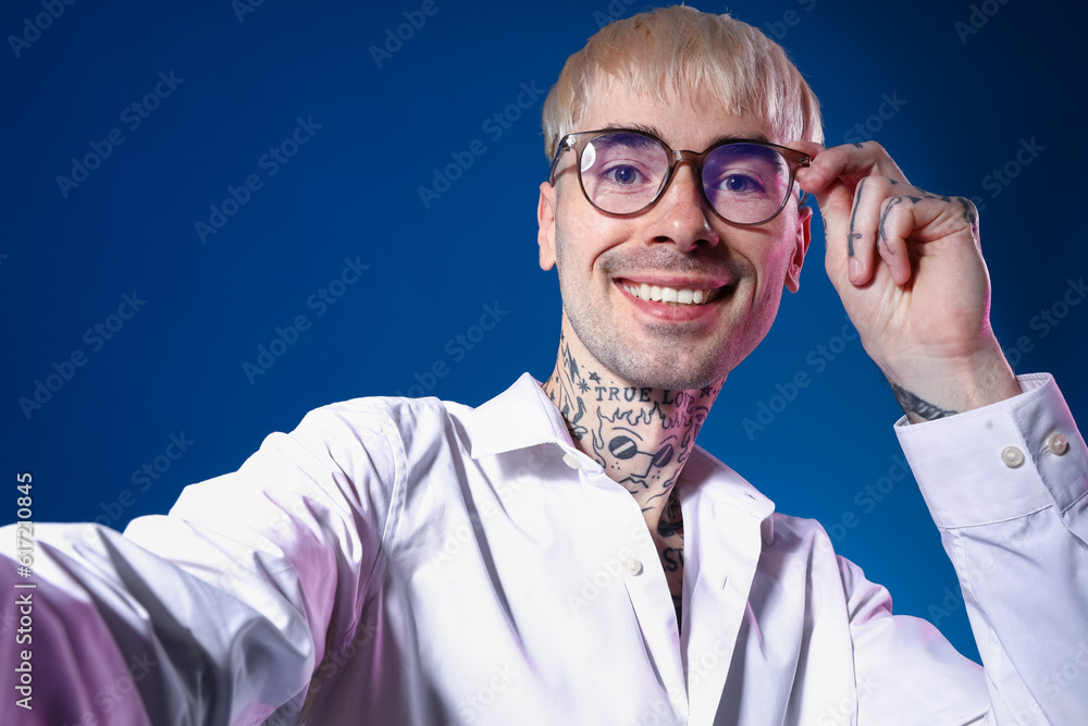 Young tattooed man in eyeglasses taking selfie on blue background, closeup