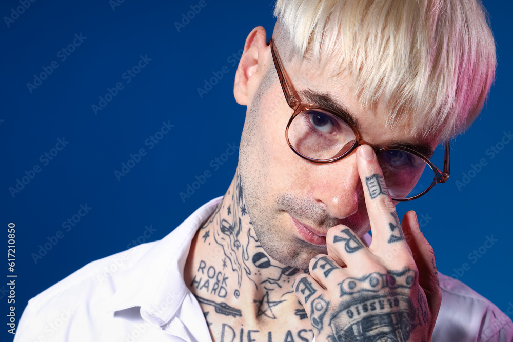 Young tattooed man in eyeglasses on blue background, closeup