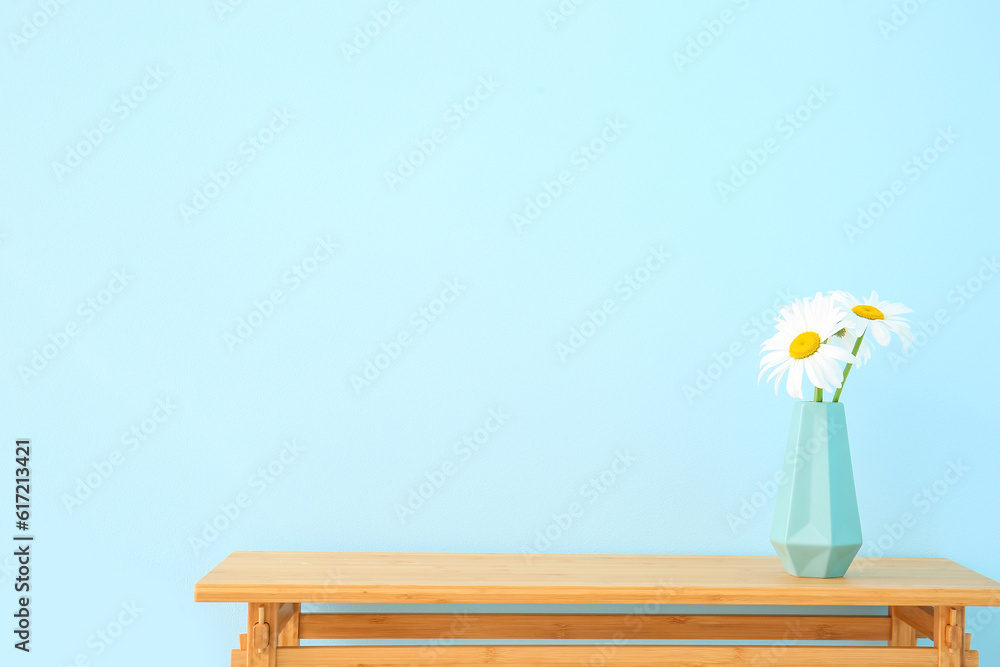 Wooden table with blooming daisy flowers in vase near blue wall