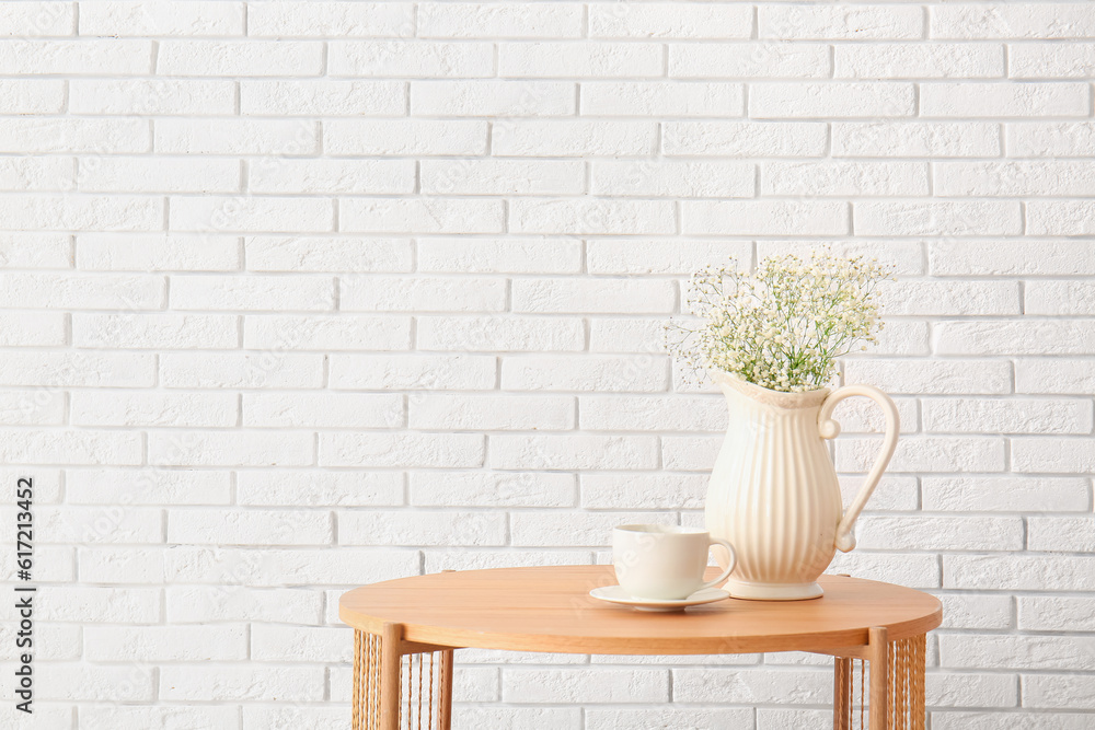 Vase with beautiful flowers and cup on wooden coffee table near white brick wall