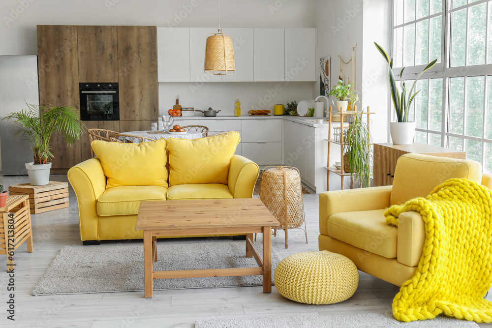 Interior of light open space kitchen with yellow sofa, armchair and wooden coffee table