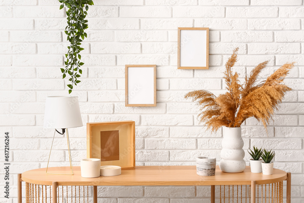 Candles, lamp, pampas grass and blank frames hanging on light brick wall in room