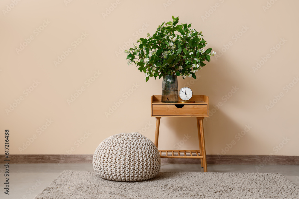 Vase with blooming jasmine flowers and alarm clock on wooden table near beige wall