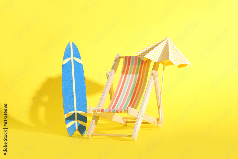 Creative composition with miniature surfboard, deckchair and umbrella on yellow background