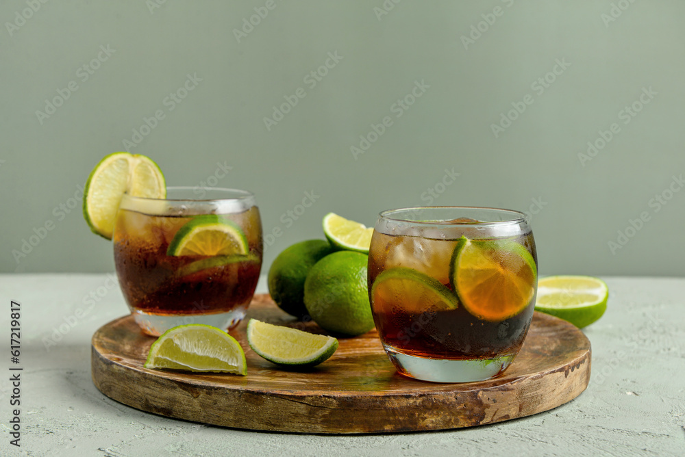 Board with glasses of cold Cuba Libre cocktail and limes on grey background