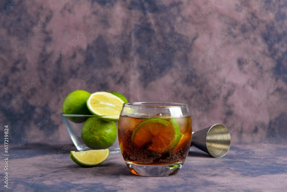 Glass of cold Cuba Libre cocktail and bowl with limes on purple background