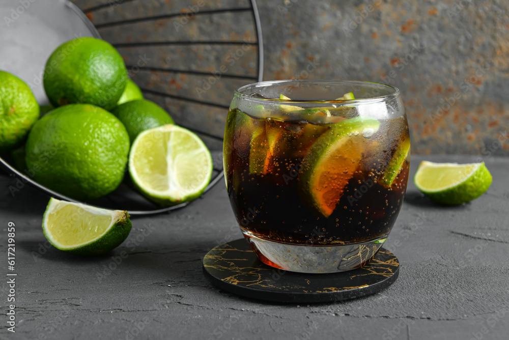 Glass of cold Cuba Libre cocktail and basket with limes on grey table