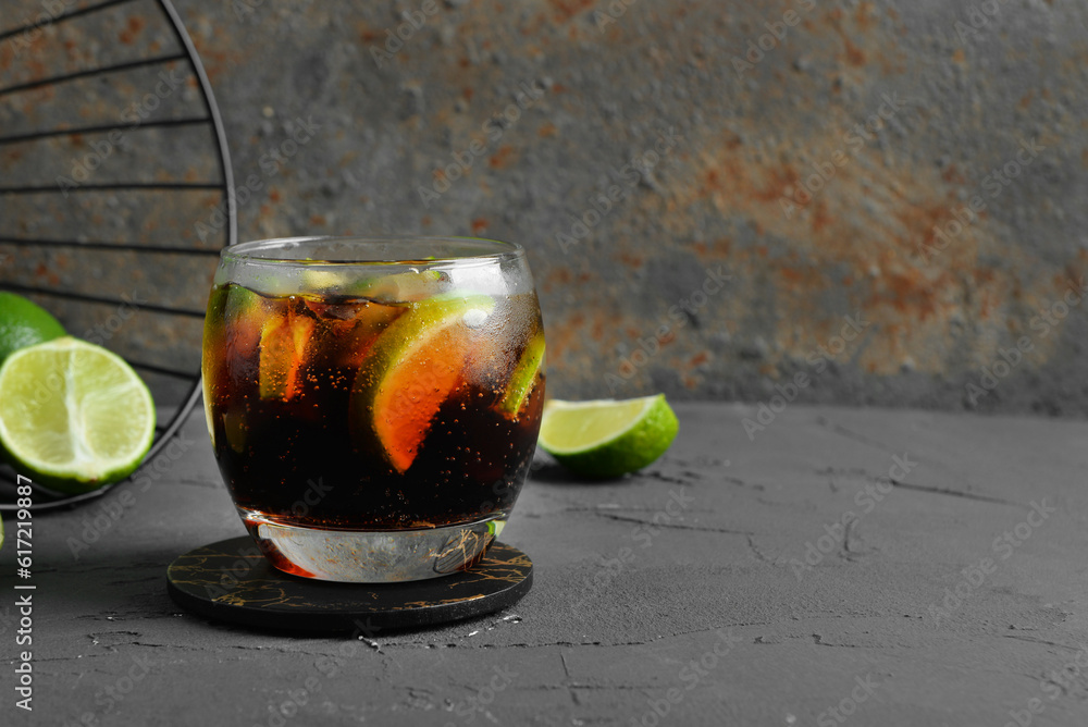 Glass of cold Cuba Libre cocktail and basket with limes on grey table