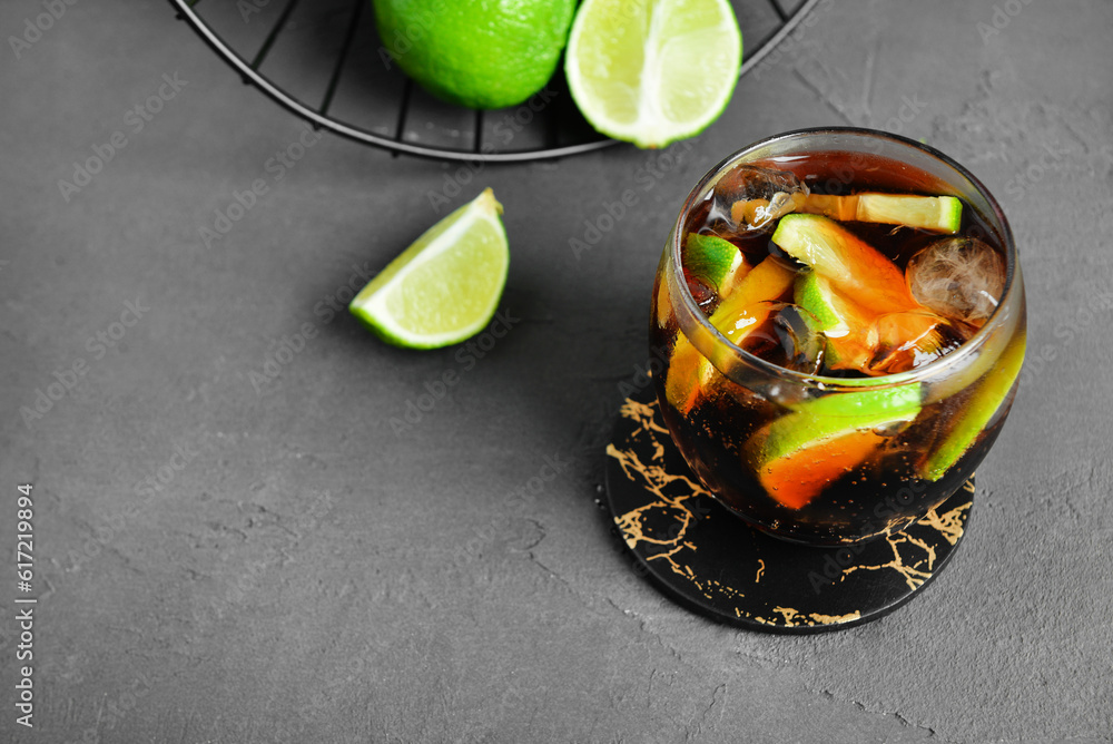 Glass of cold Cuba Libre cocktail and basket with limes on grey table