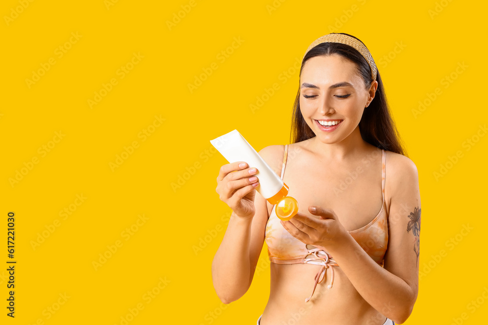 Young woman applying sunscreen cream on yellow background