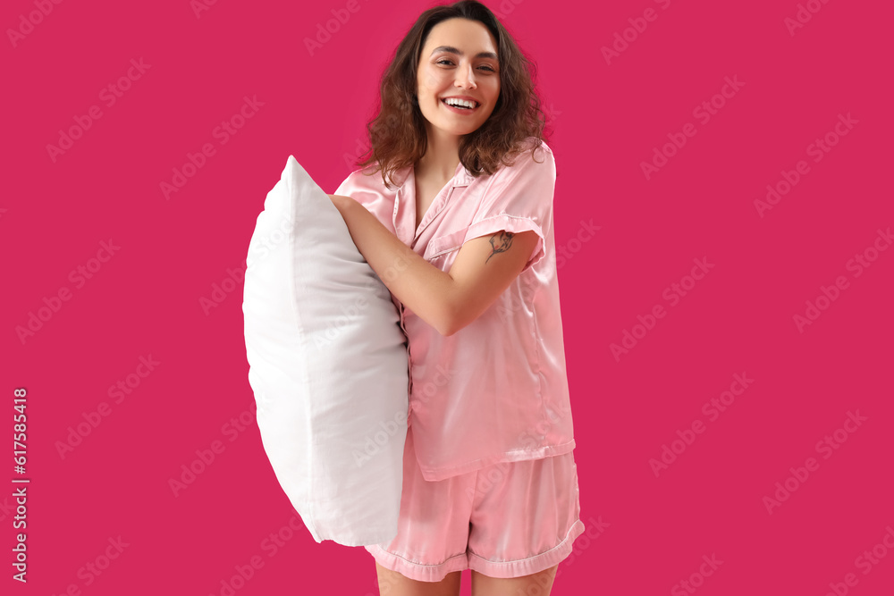 Young brunette woman in pajamas with pillow on pink background