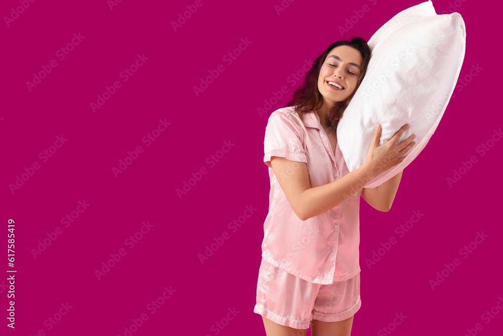 Young brunette woman in pajamas with pillow on pink background