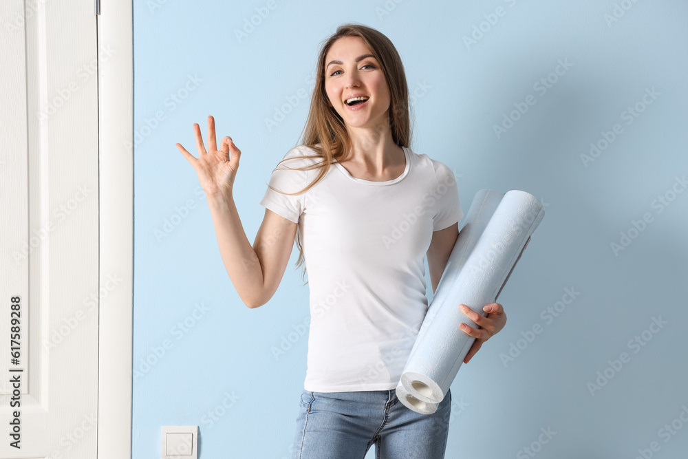 Young woman with wallpaper rolls showing OK near blue wall