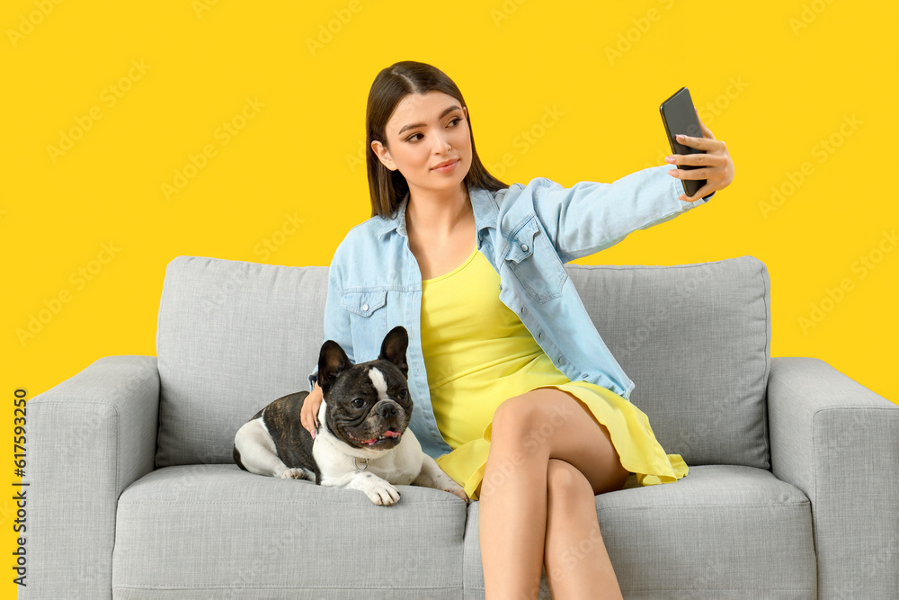 Young woman with her French bulldog taking selfie on sofa against yellow background