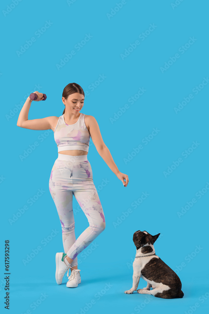 Sporty young woman with dumbbell and her French bulldog on blue background