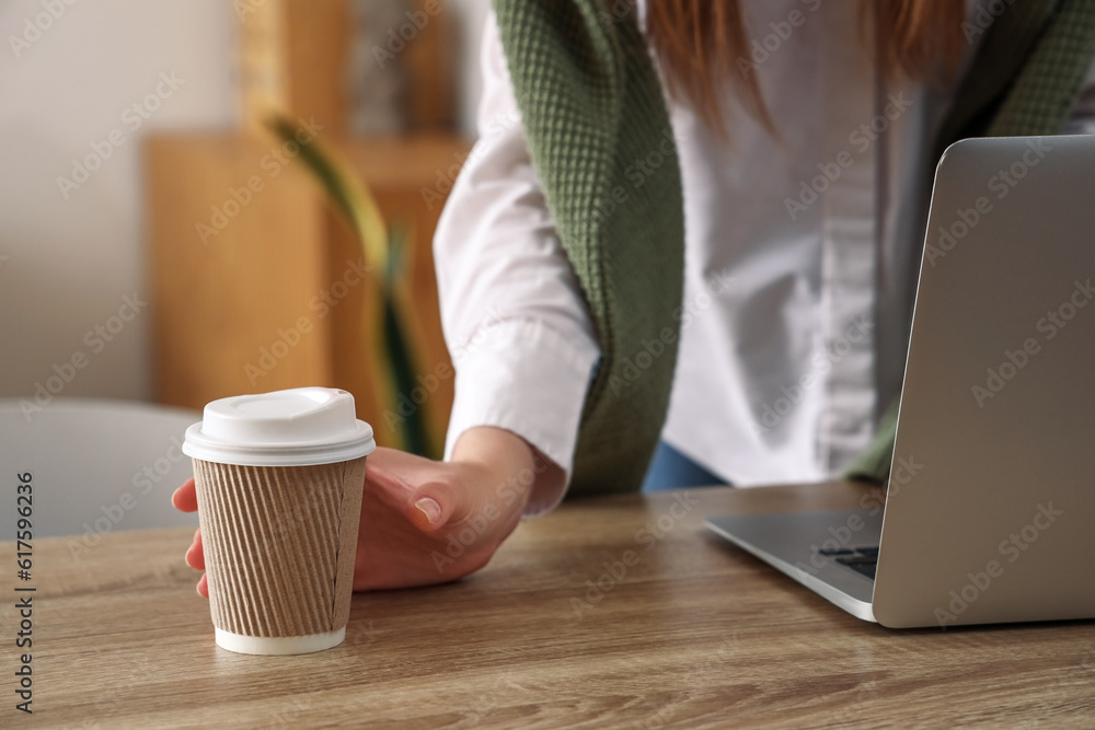 Woman using modern laptop and holding takeaway cup of coffee at table