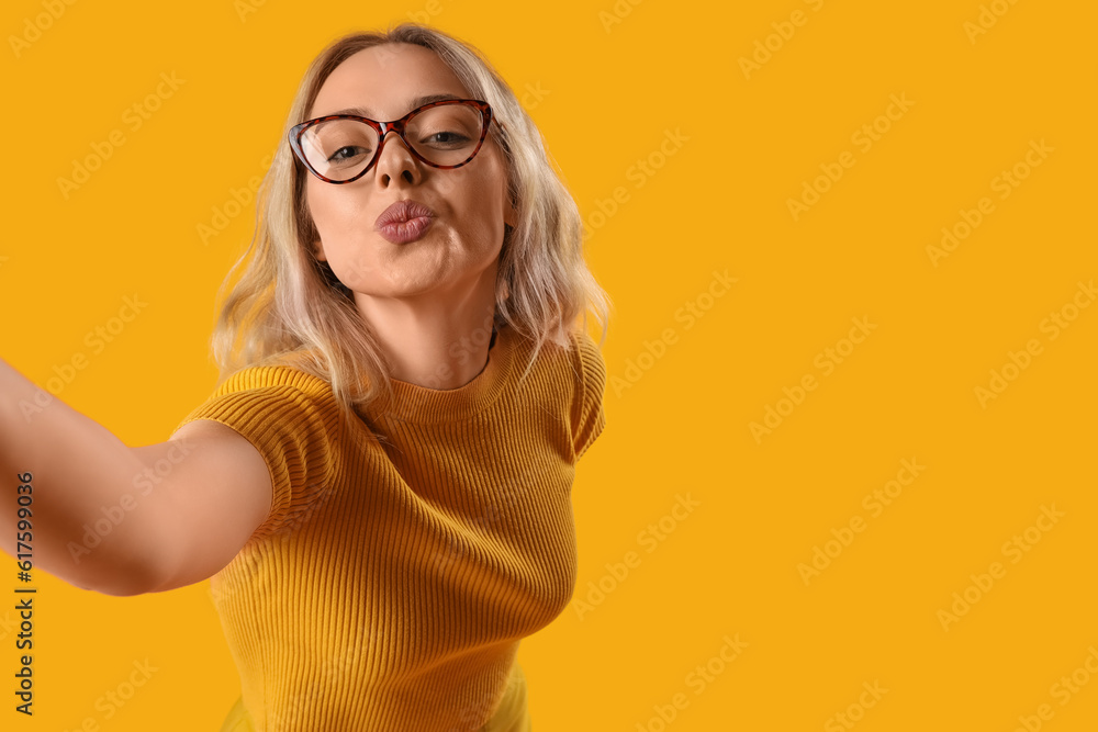 Young woman in stylish eyeglasses blowing kiss on orange background, closeup