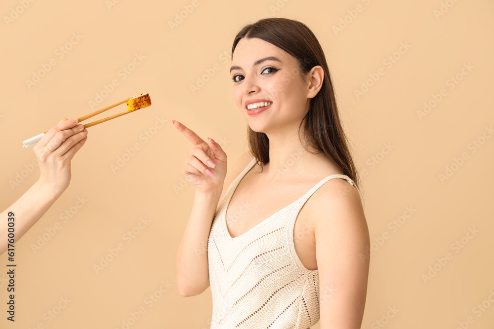 Young woman pointing at female hand with sushi roll on beige background