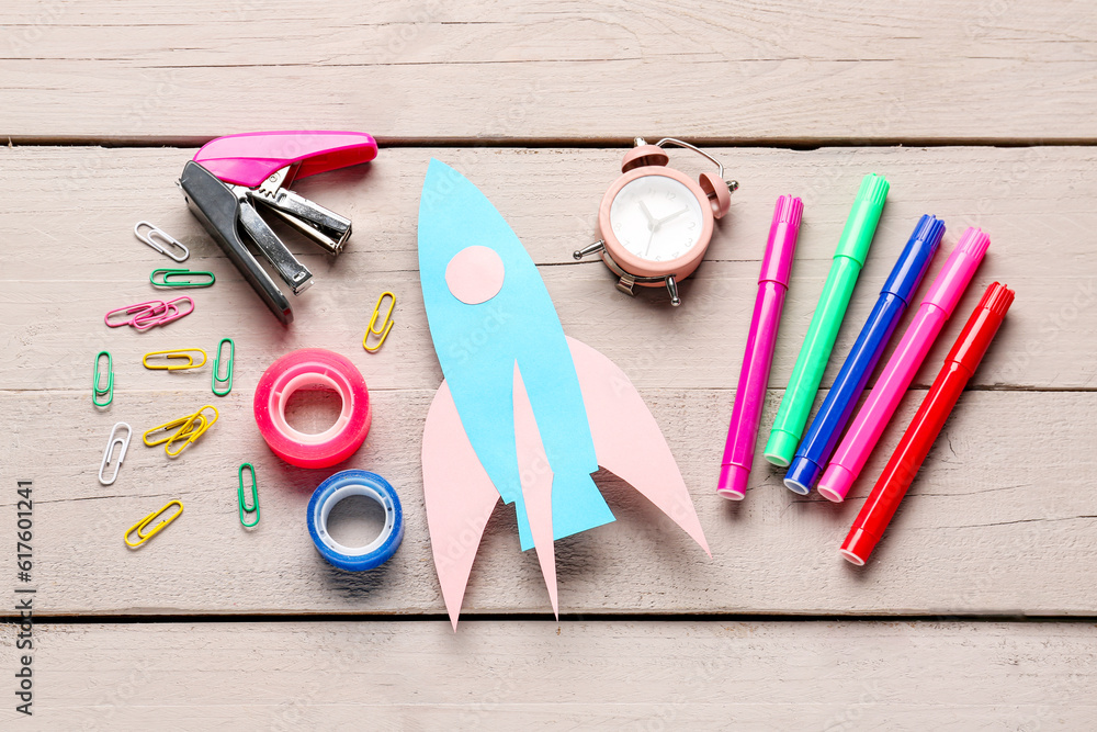 Blue paper rocket with alarm clock, markers and stapler on beige wooden background