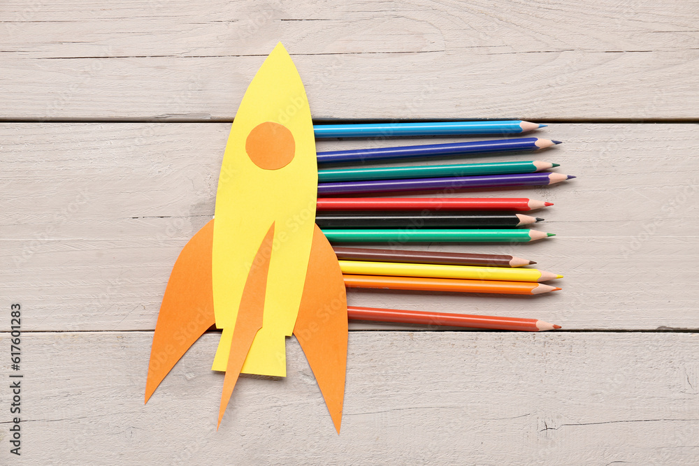 Yellow paper rocket with color pencils on beige wooden background